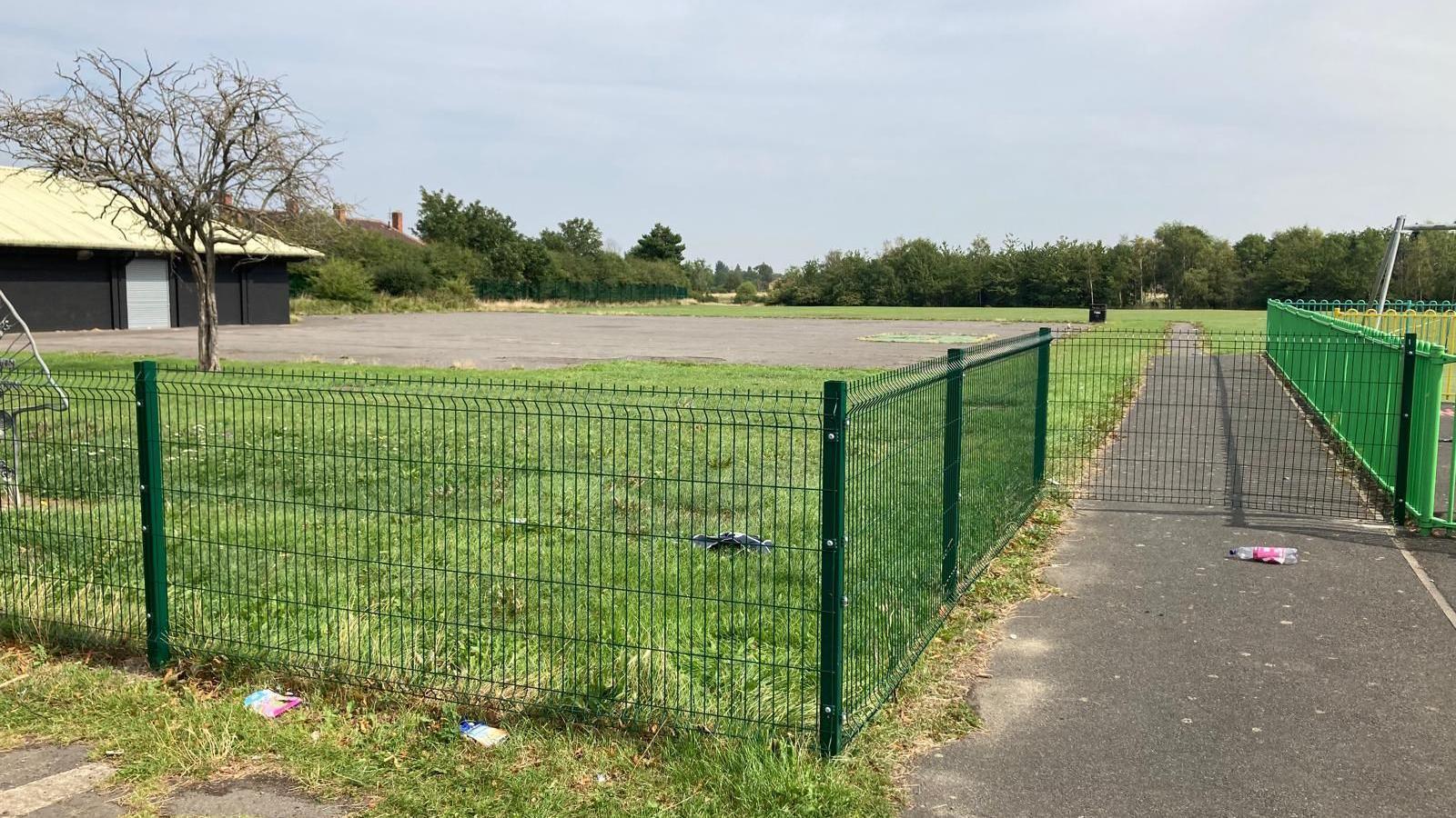 A green wire fence surrounds a field which is a mix of grass and concrete. There is a single-storey hut on the left and trees in the distance.