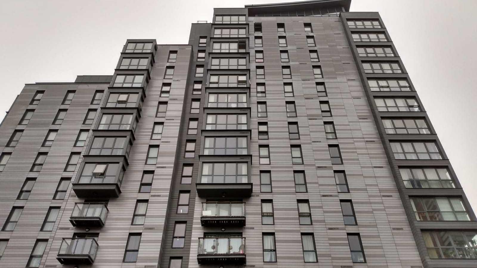 A photo of the grey and black tower block taken from the ground next to it, with the tower looming over the photographer against a white cloudy sky.