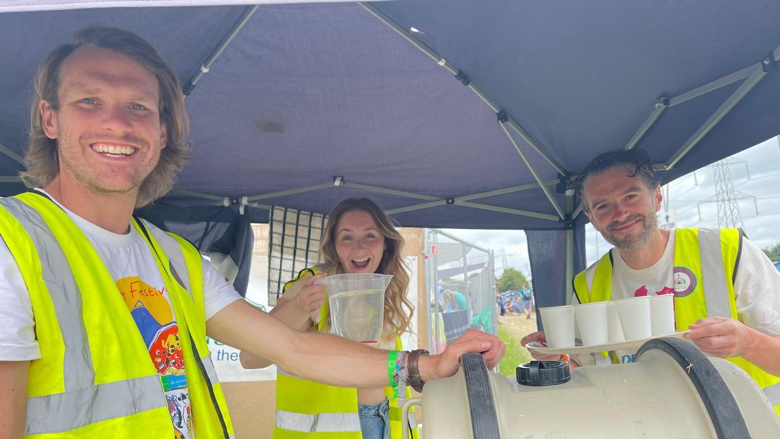 Three people, in high-vis jackets, handing out water. Lewis, left, has on a white patterned T-shirt, a woman is holding a water jug and a man is holding a tray full of cups. They are by a water barrel.