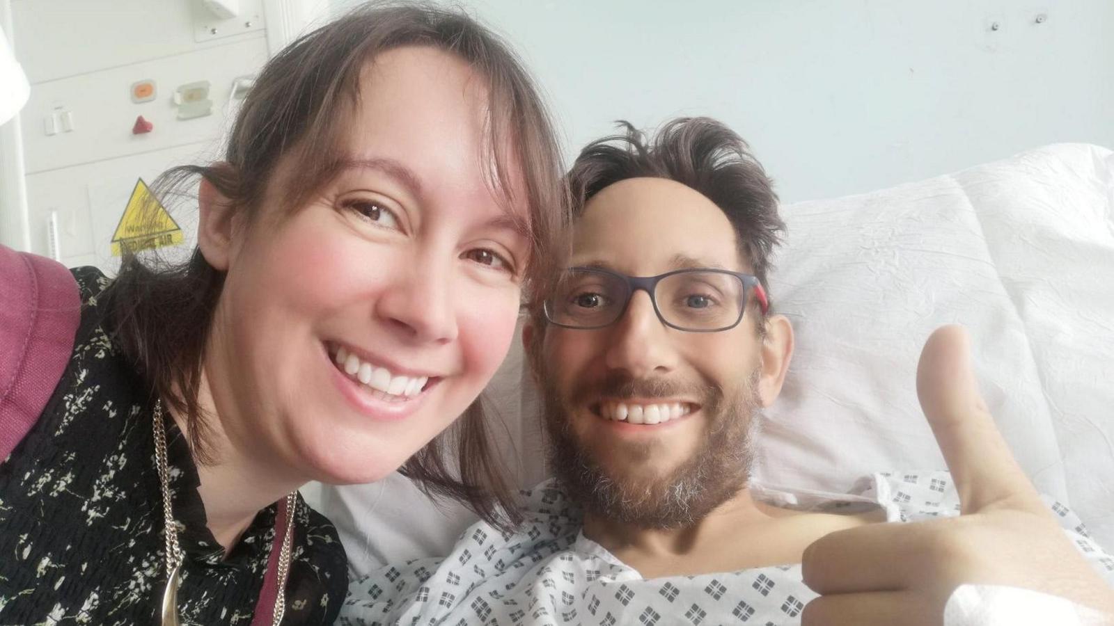 James Lawton in a hospital bed wearing a gown putting a thumb up for the camera, with his wife leaning over him to get into the picture.