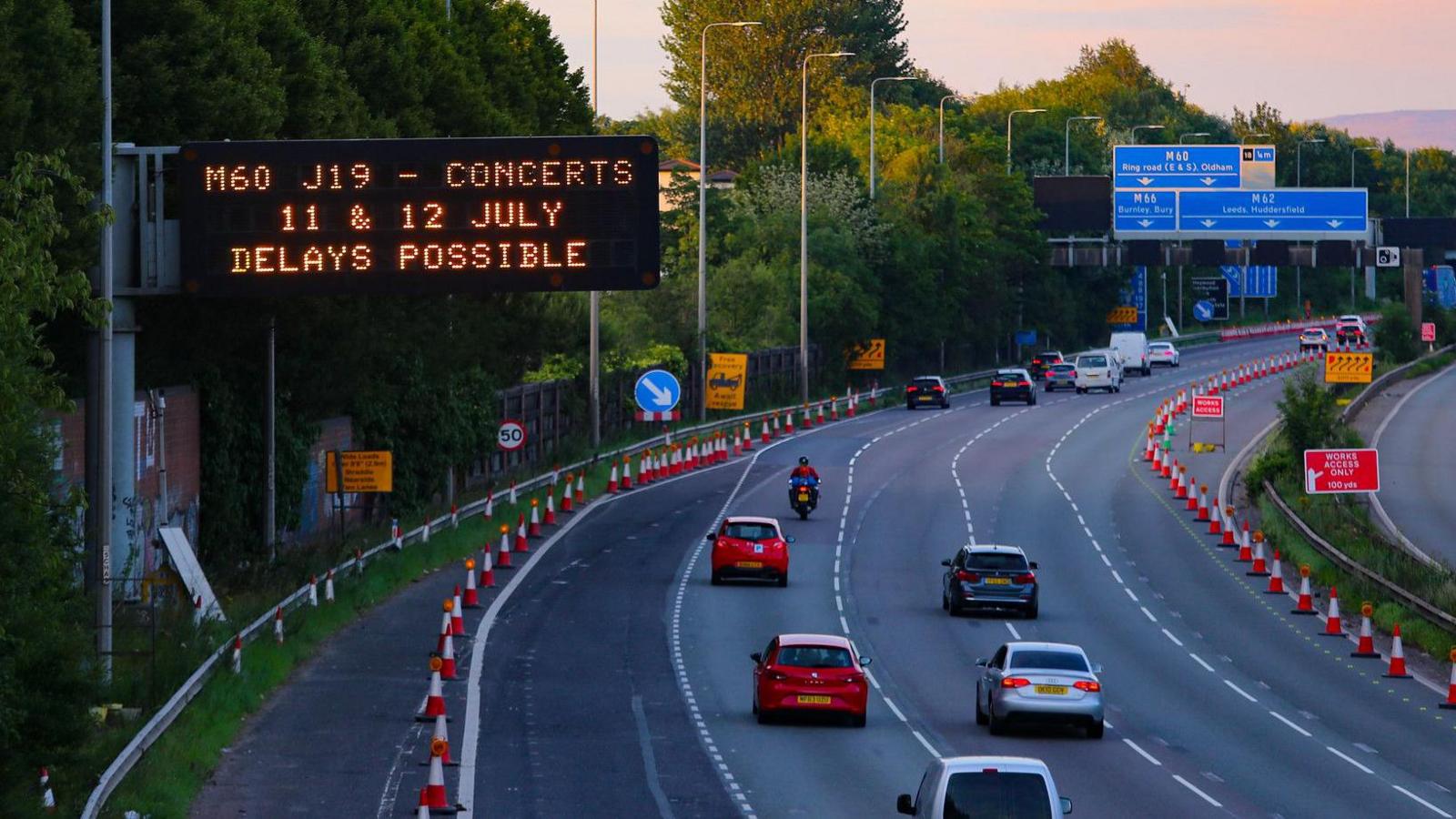 A motorway sign above a carriageway, which has a few cars driving past into the distance, reads: M60 J19 - Concerts 11 & 12 July Delays Possible