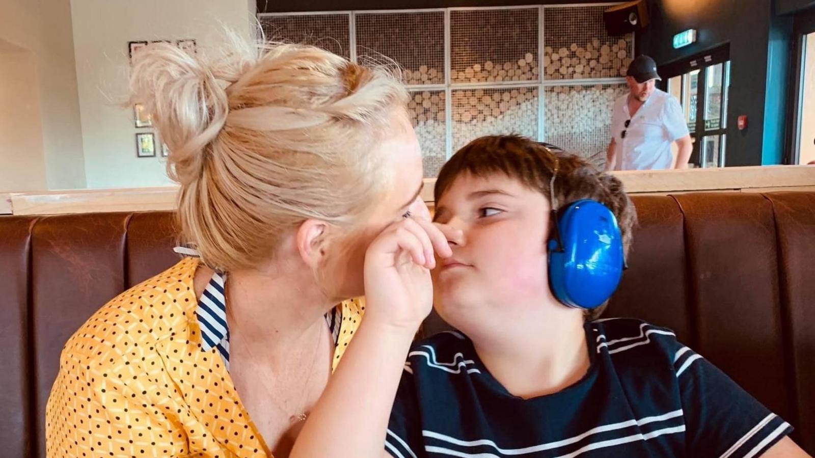 A closeup of Charlotte and Tobie sitting together. They are looking into each other's eyes in a restaurant. She is wearing a  yellow blouse. He is wearing a blue and white stripy t-shirt and blue ear protectors. 