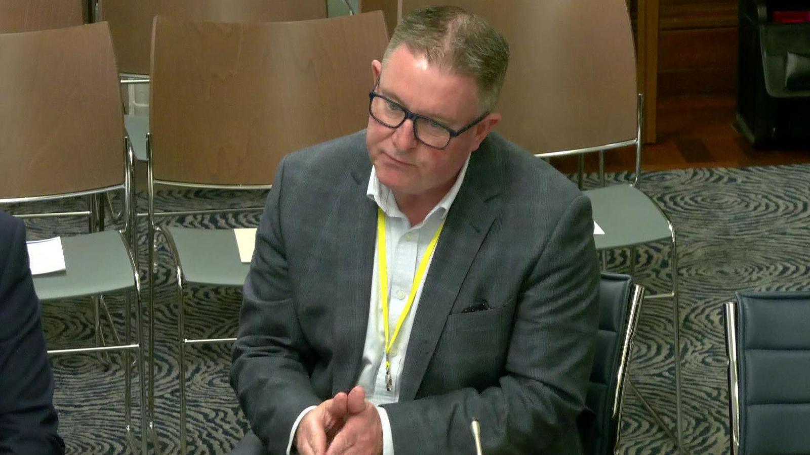 A man is sitting in a room. He wears glasses and has a grey suit on with a white shirt. He has a yellow lanyard on. There are empty chairs behind him.