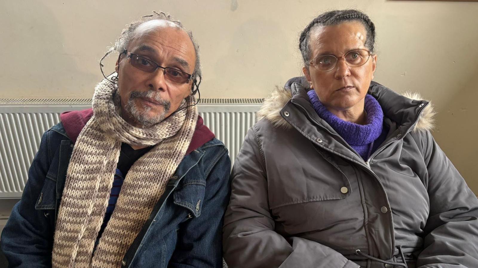 A man and woman are sitting in front of a cream wall. There is a radiator behind them and they are both looking into the camera. The man is wearing rectangular tinted glasses, a cream scarf and a denim jacket. The woman is wearing a purple turtleneck jumper and a grey padded coat with fur around the hood.