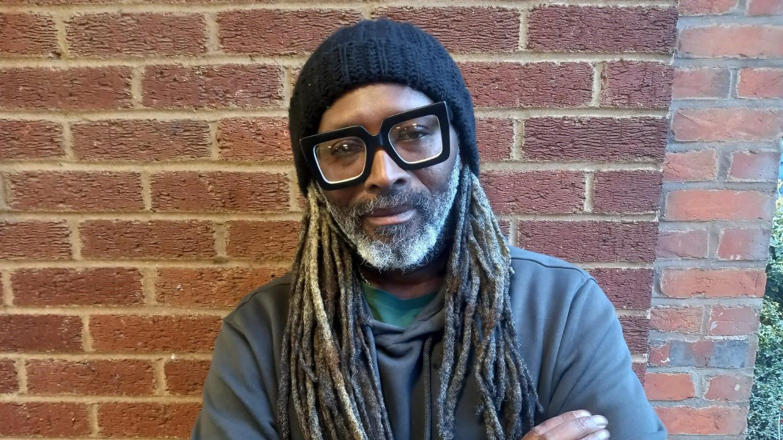 Lindsay Dube. He has long dark hair, wearing a dark grey jumper, black beanie and glasses. A red brick wall is behind him.