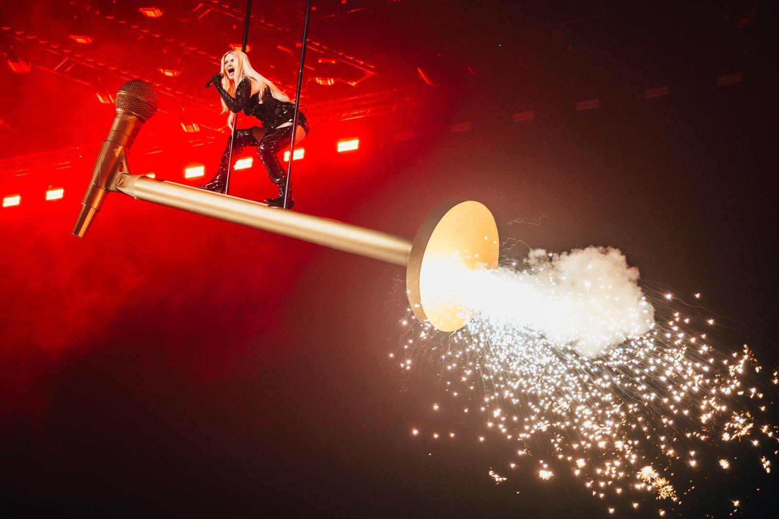 Erika Vikman stands on a giant gold prop microphone, as fireworks explode around her