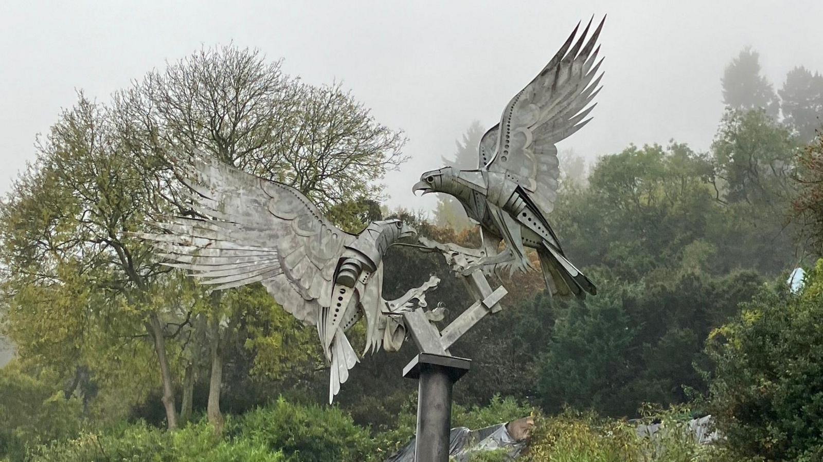 A metal sculpture of two birds fighting with their wings outstretched at the top of a metal pole with green leaved trees behind them.
