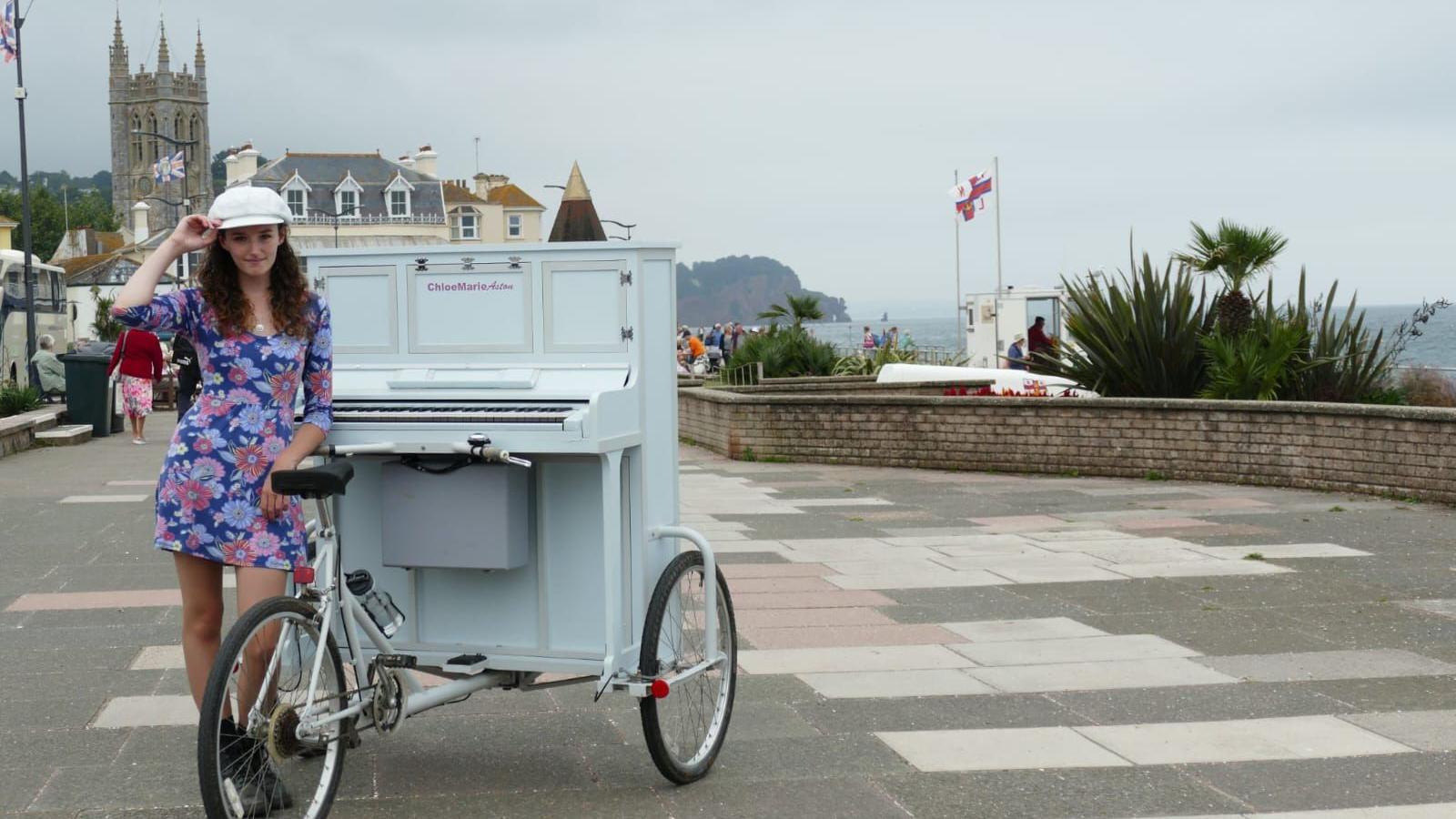 Chloe Marie Aston is seen with her piano bike inTeignmouth, Devon. She is wearing a short pink and blue patterned dress. The town can be seen behind her with St Peter's Church in the background.