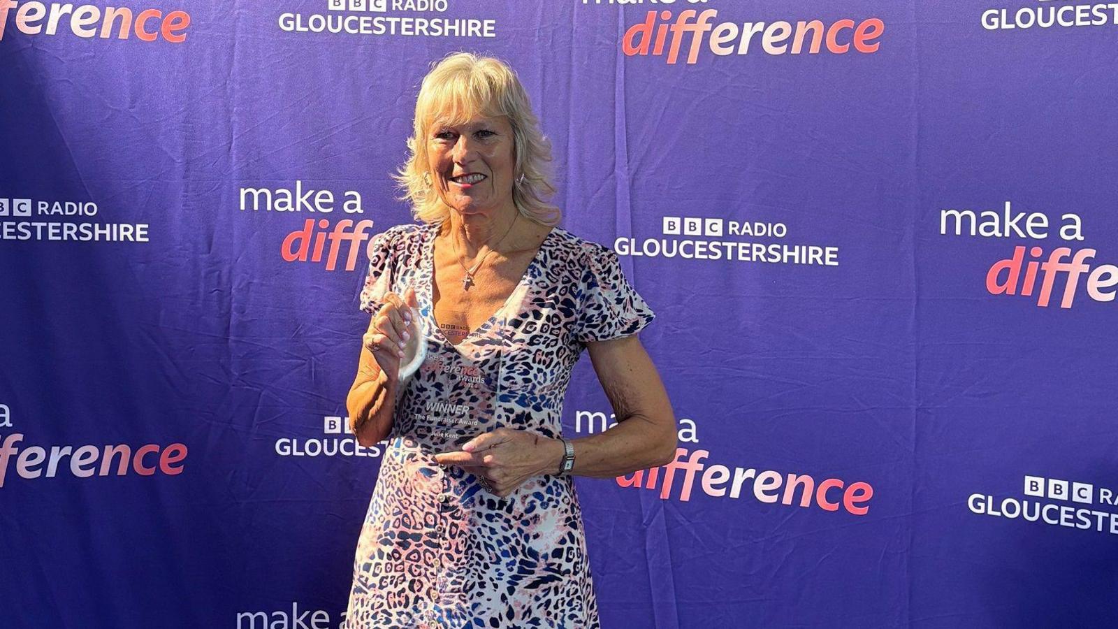 Julie Kent holding her Make a Difference Award. She's wearing an animal print dress and is standing in front of the Make A Difference branding.