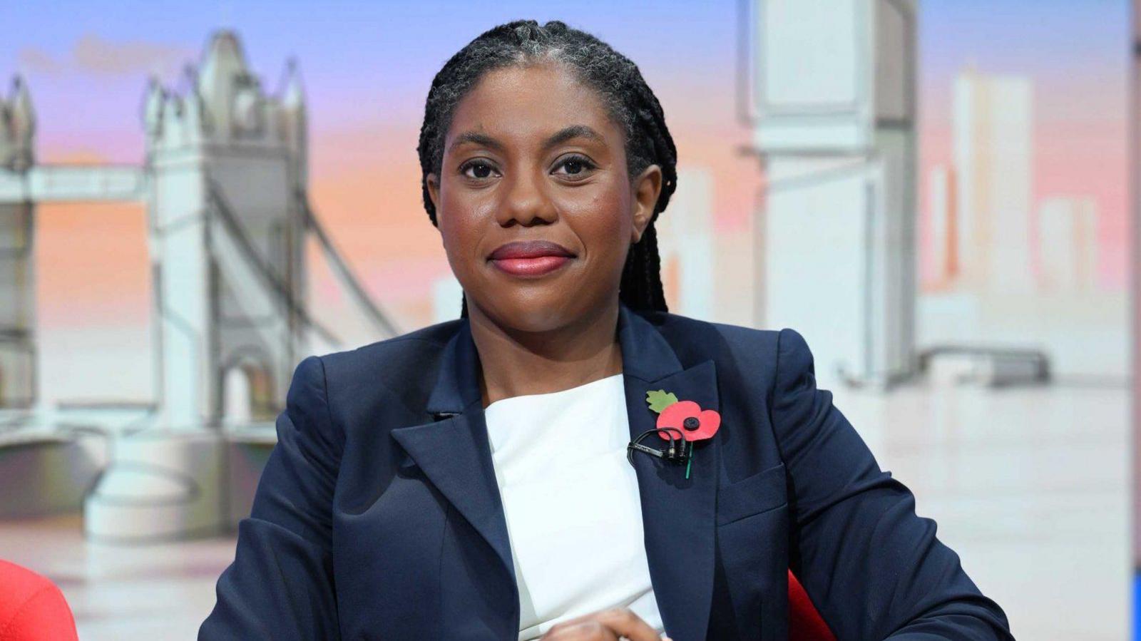 A woman wearing a dark blue blazer and a white top sits in front of an animated bridge. She has a poppy on her lapel. 