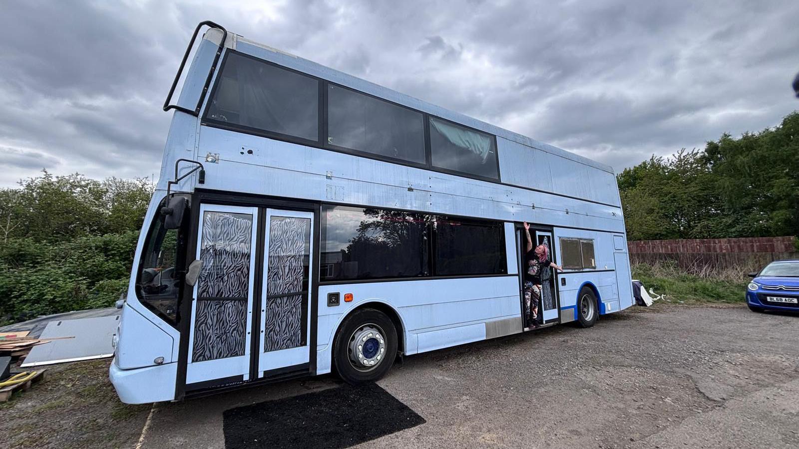 Lyndzi Hargrave smiling with her arms outstretched leaning out of the side of a double decker bus with a light blue exterior.