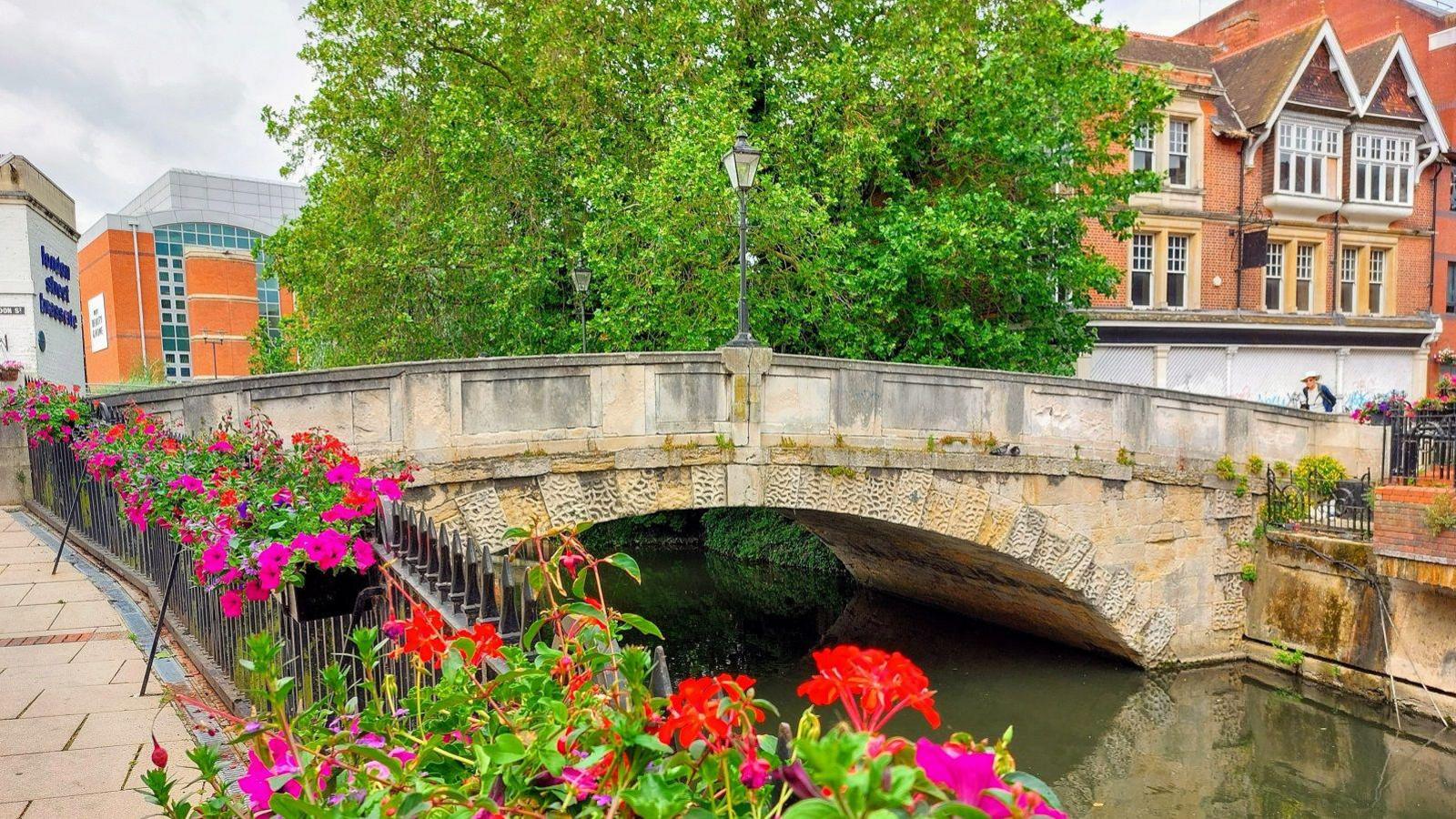 An old stone bridge going across a river, with red and pink flowers in the foreground and a tree in the background