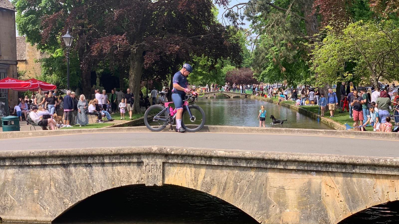 A man crosses a small, low, Cotswold stone bridge over the shallow River Windrush on a sunny day. On either side of the river, tourists can be seen standing on the grassy banks, with a girl and a black dog playing in the water. There are trees hanging over the river.