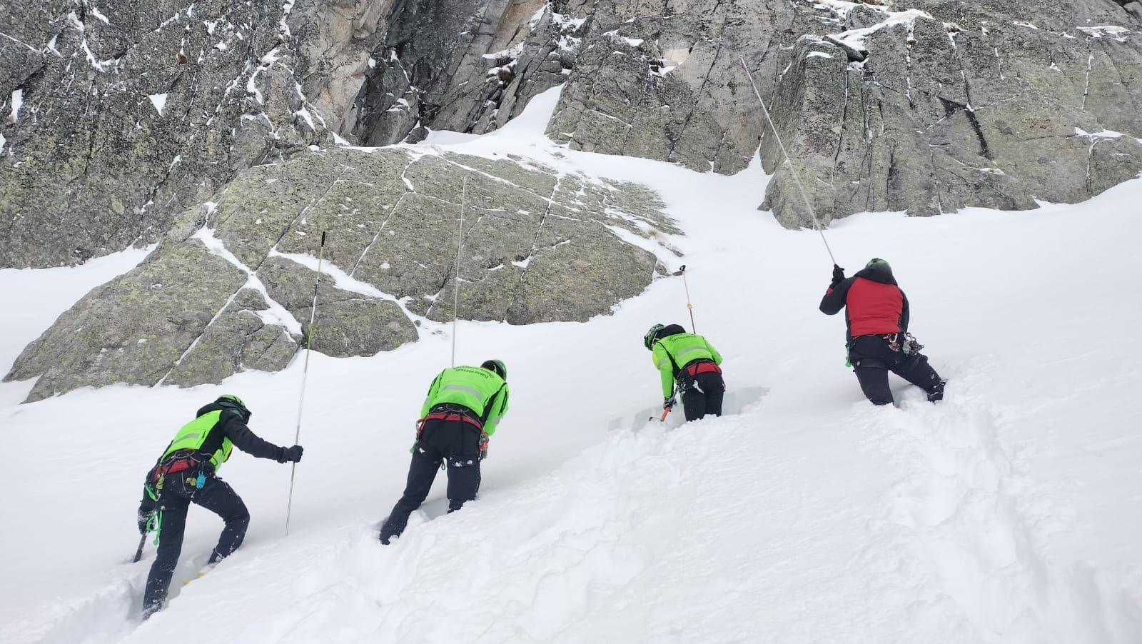 Four alpine rescue search team members wearing high vis uniforms search the snowy side of a mountain with ice picks and long poles.