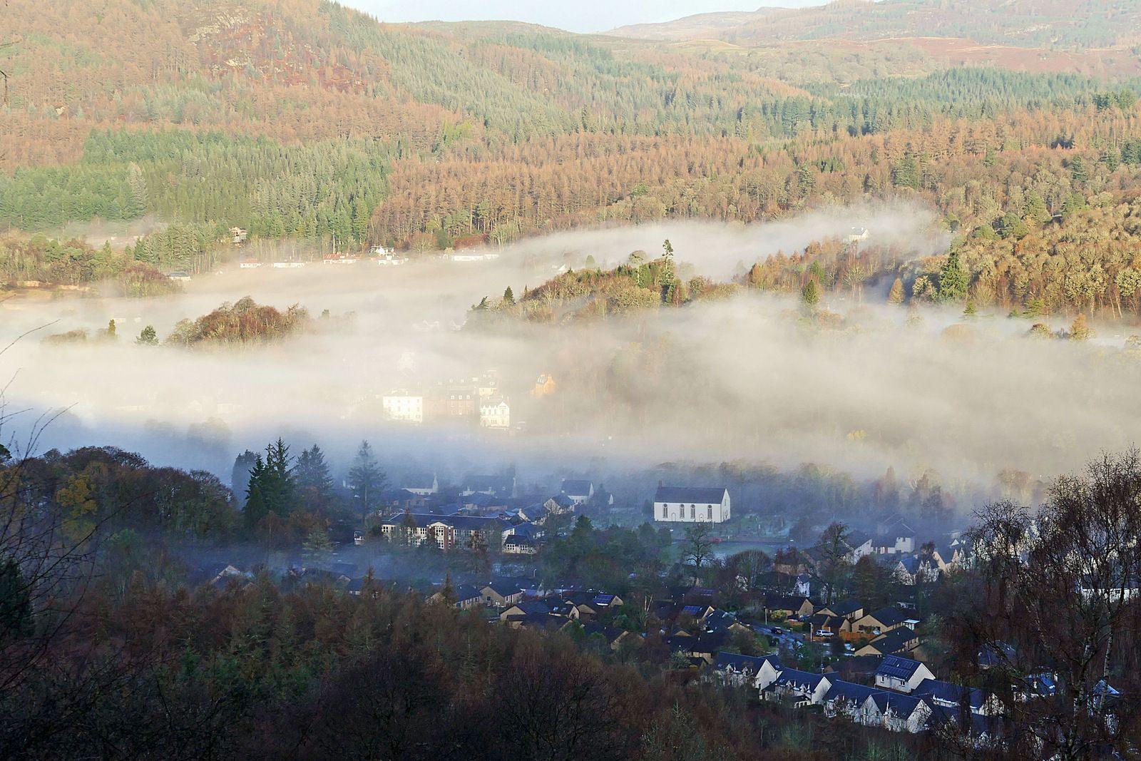 Overhead image of a small village nestled among hills and a tree-covered area with low hanging fog over the houses obscuring them slightly