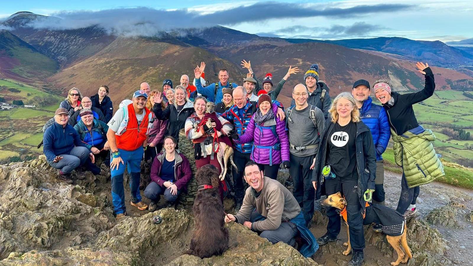 A group of walkers and dogs smile and wave at the camera at the top of a mountain in Cumbria  