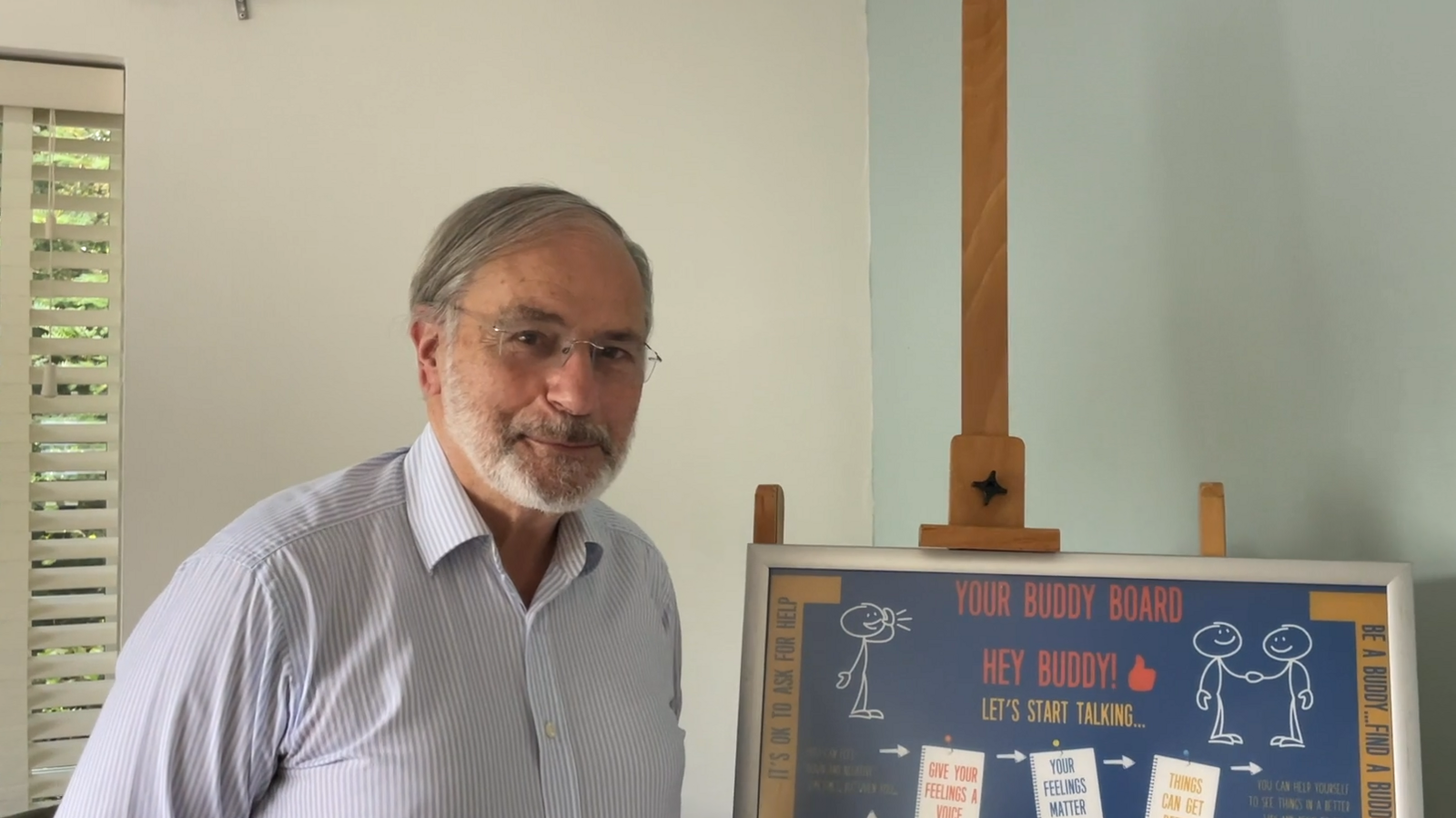 Gentleman with a white and light blue striped business shirt on, glasses and a grey beard and hair, next to a "Buddy Board" for his charity Joe's Buddy Line.