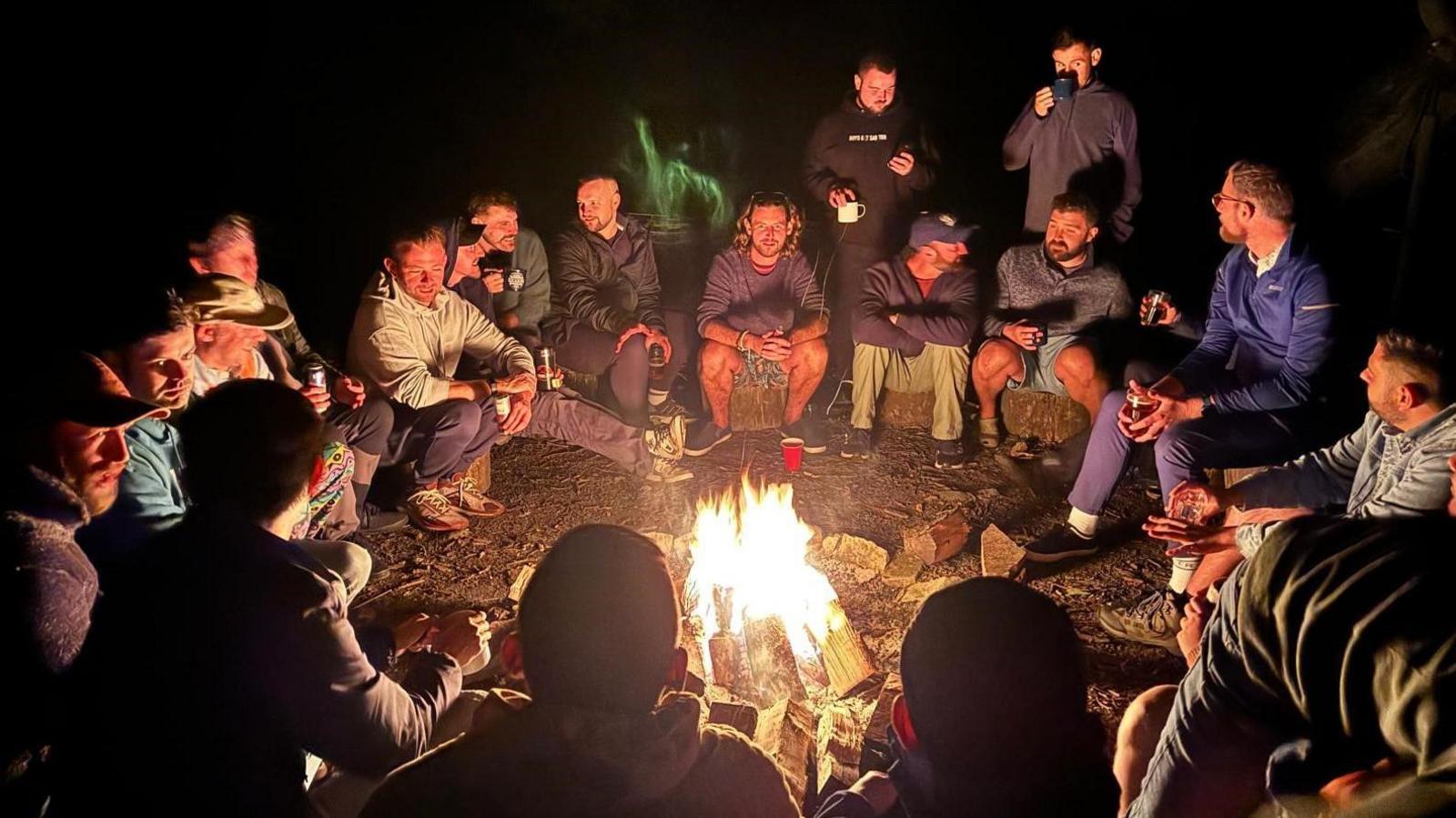 A group of men sat around a camp fire at night. Several of them are holding cups.