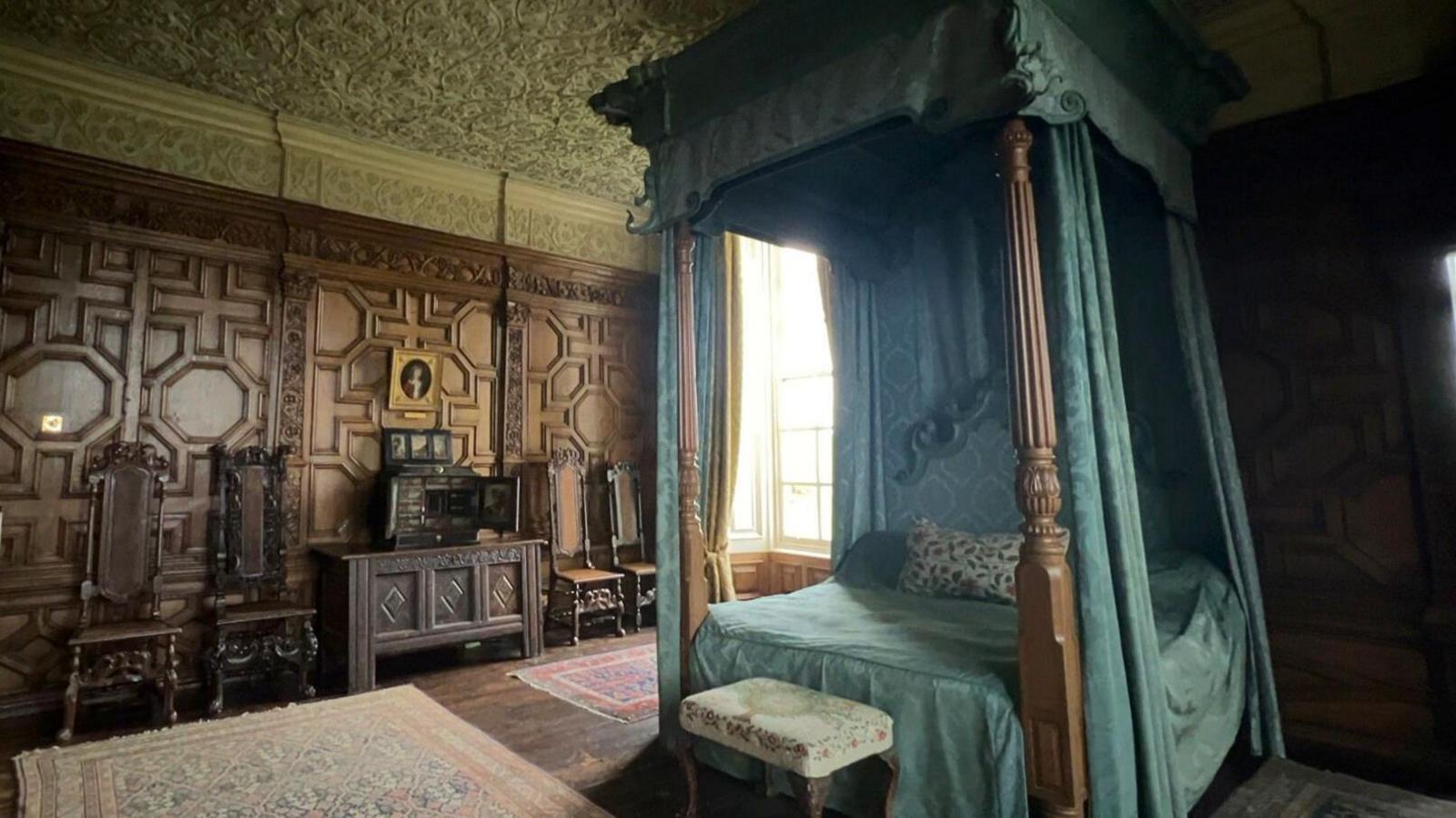The Queen's state bedroom at Burton Agnes Hall. There is a large, wooden four-poster bed in the centre of the room, it has blue curtains and bedding. The walls are wood panelled in a geometric pattern. To the left of the bed, there are four wooden, high-backed chairs and a wooden dresser. There is a gold-framed painting on the wall of a boy in a red shirt.