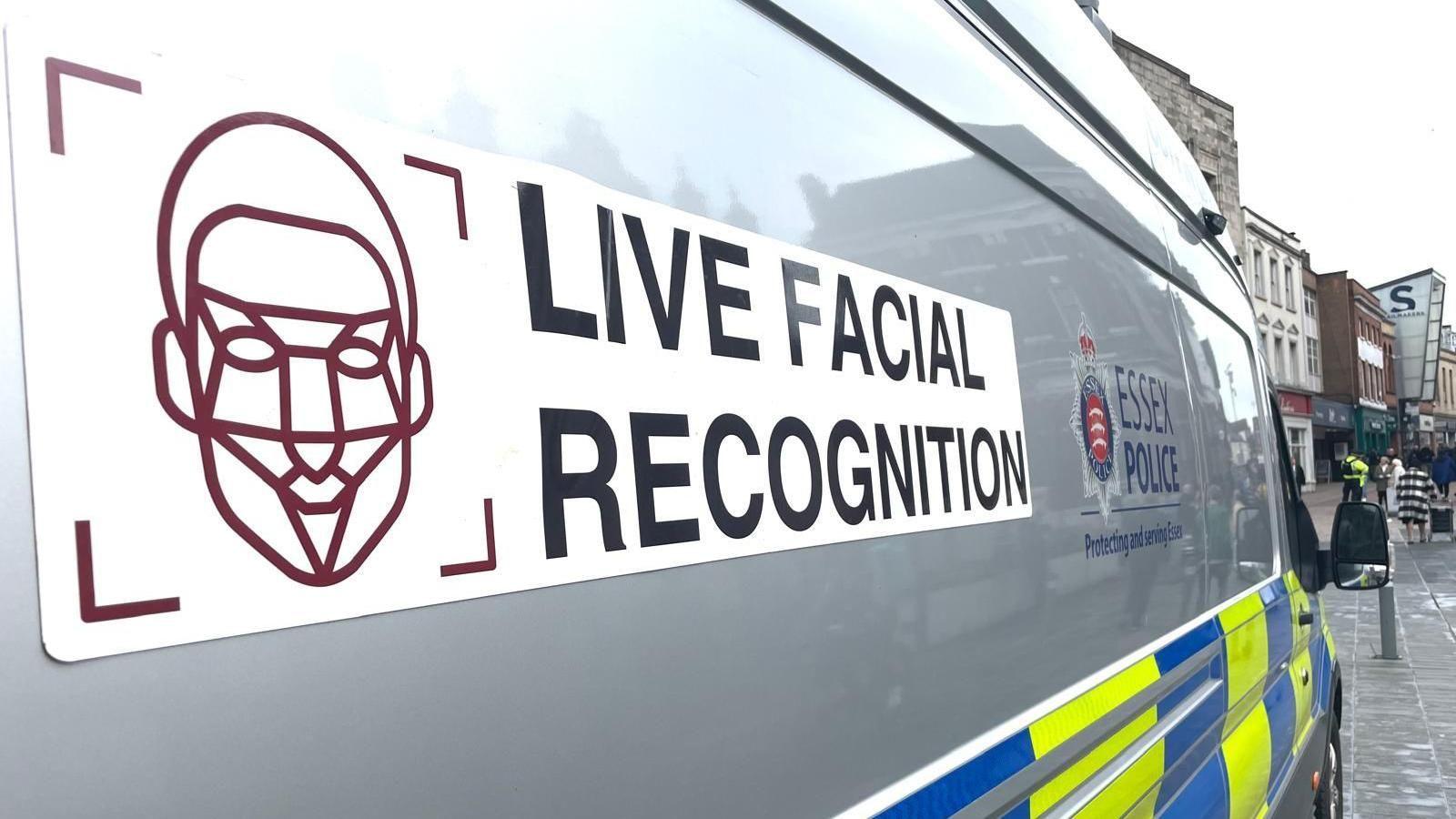 A view taken to show the side of a facial recognition police van. Closest to the camera is a stylised graphic of a face being scanned, next to the text 'Live Facial Recognition'. Further along the van, only partially visible, is the logo for Essex Police