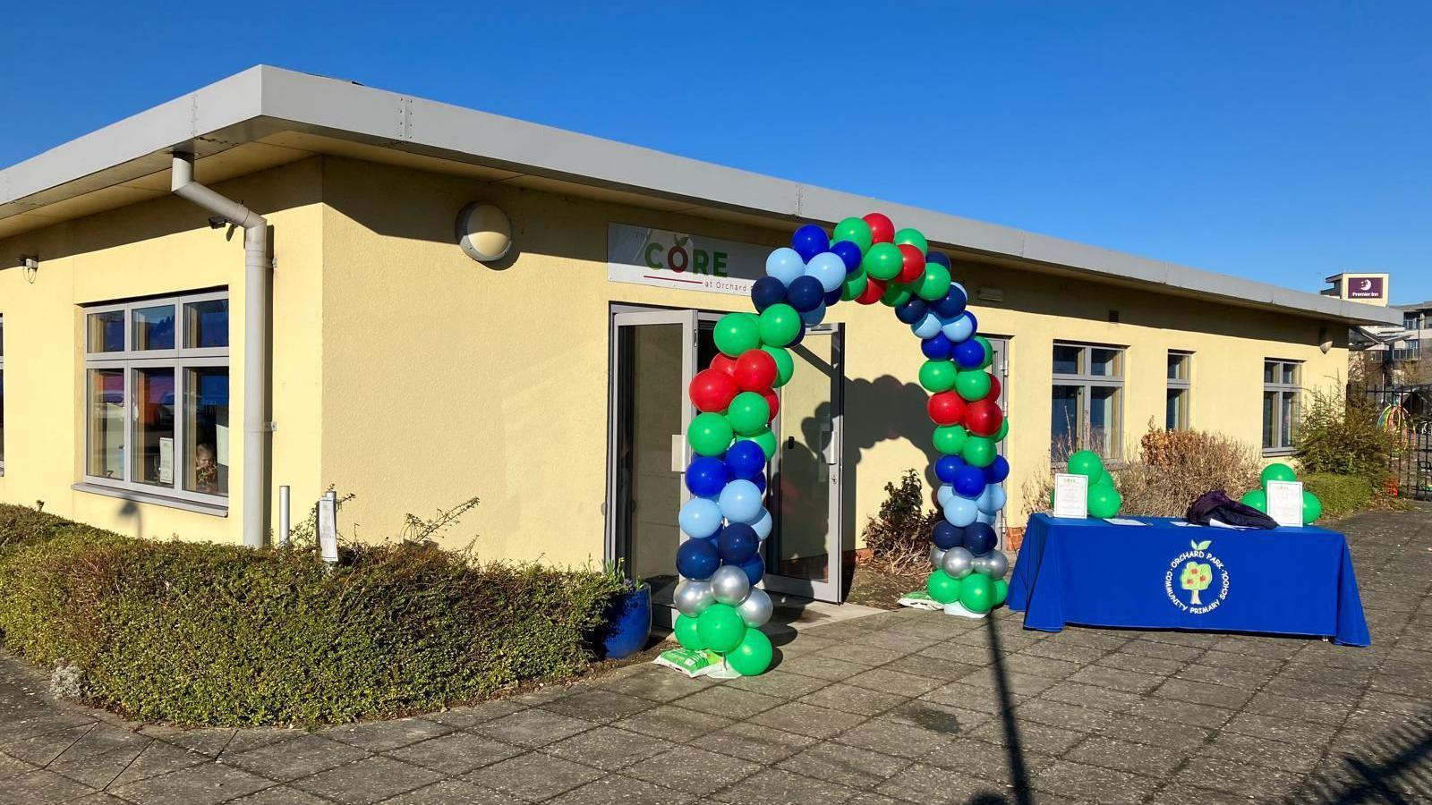 The outside of the community hub. It is a yellow single-storey building, with an arch of coloured balloons by the entrance.