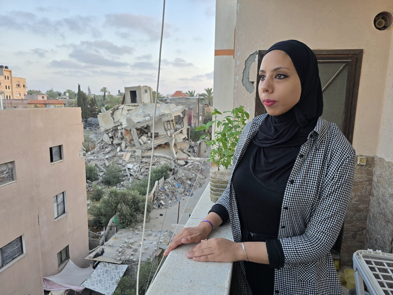 Hanya, who is wearing a black headscarf, looks out at a street filled with destroyed buildings, from her balcony