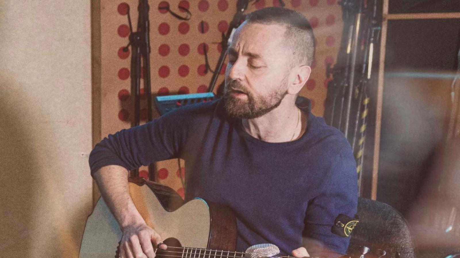 Ross Wilson sits playing an acoustic guitar. He is wearing a blue jumper and has a beard.