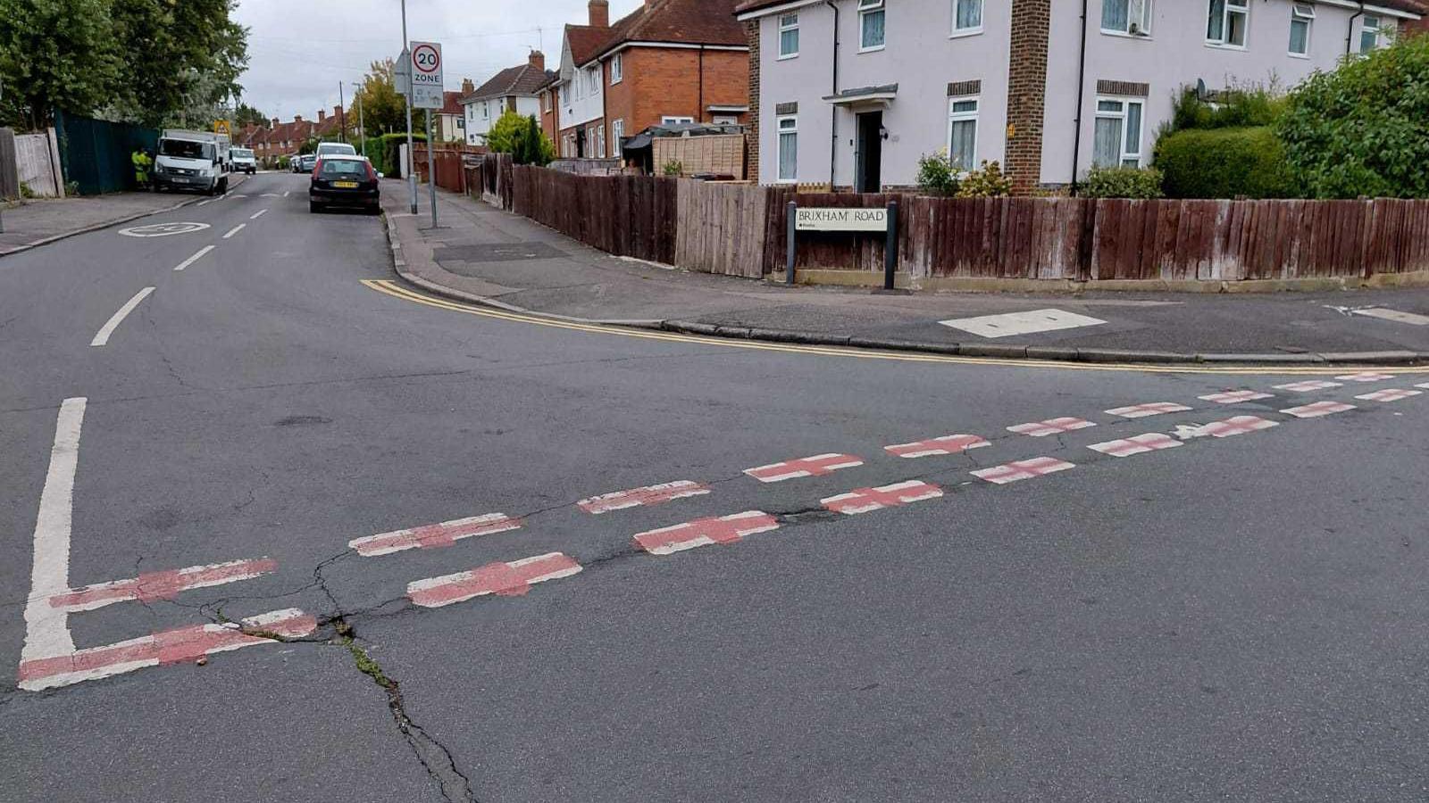 A road junction in a residential area on a cloudy day. The white "give way" dashes are each graffitied with red crosses.