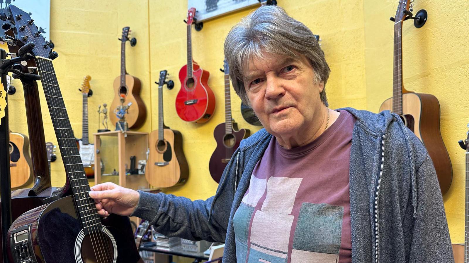 A man, wearing a grey jacket and a purple shirt, with his right hand on the fretboard of a dark-coloured acoustic guitar. Behind him is a yellow wall with guitars mounted on it.