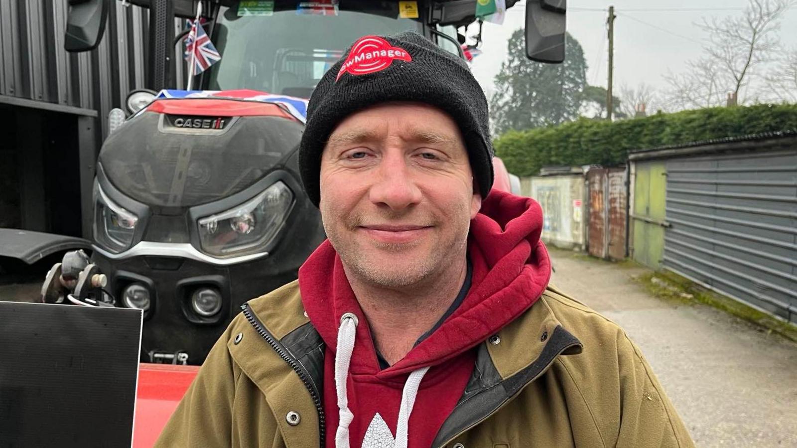 A man in a black beanie, red hoodie and green coat. He is standing in front of a camera.