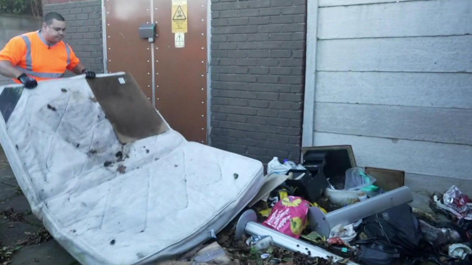 A man in an illuminous orange hi-vis top lifting a mattress which is among the illegal rubbish which has been dumped in Sefton.