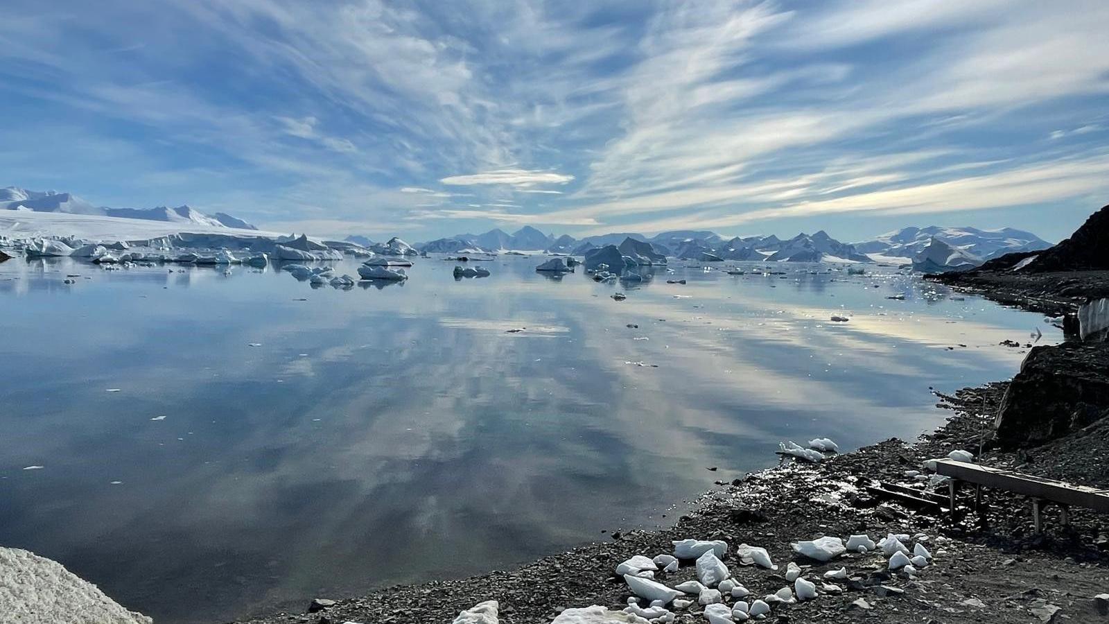 A landscape photo of water across Antarctica taken from a shore. The sky is reflected in the water and there are icebergs and mountains in the distance.