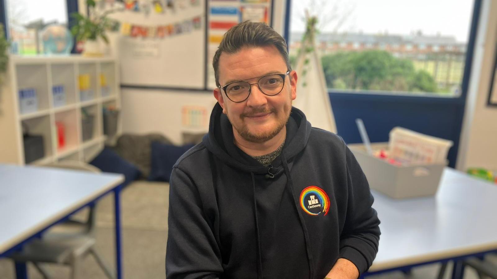 Stuart Mycroft smiles into the camera in a classroom at his school. Desks and a cushioned floor seating area are blurred in the background. Mr Mycroft is wearing glasses and a dark hoodie with a rainbow emblem on the front.