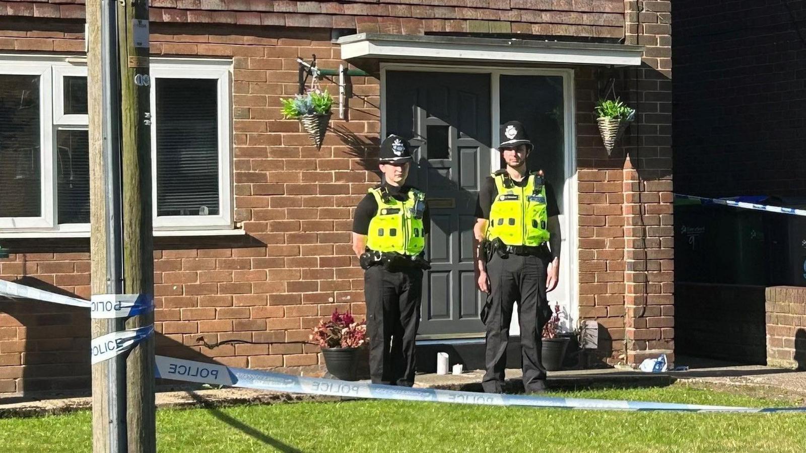 Police tape around a house, with two police officers stood outside the front door