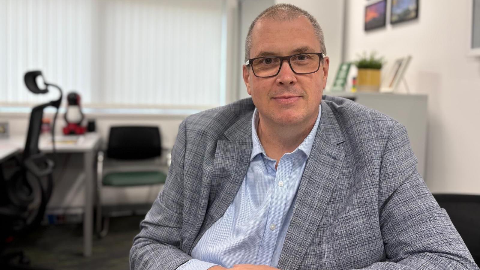 Andy is wearing a shirt and jacket and is sat in an office. He is wearing dark-rimmed glasses and has a neutral expression.