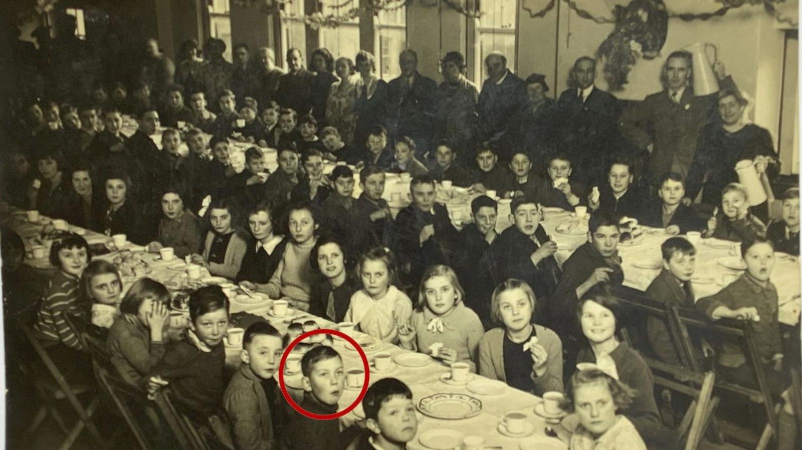 A black and white picture of children sitting at two tables in Alcombe. They are eating food. Adults are standing infront of windows in the background.