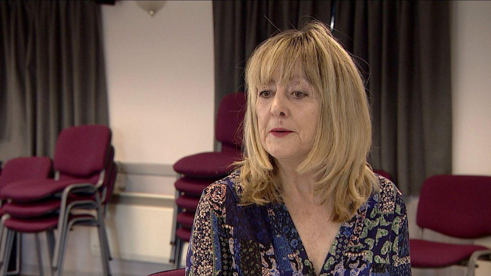 A photo of Dale Povey, a woman with shoulder-length blonde hair. She wears a blue patterned top and sits in a room where red checks are stacked behind her.