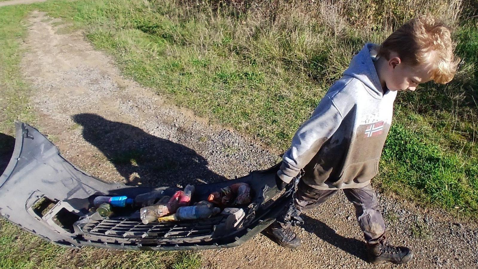 Logan Currie, 7, pulling the front bumper of a car with bottles and cans resting on it. Logan's grey hoodie is wet with mud and he is wearing small walking boots.
