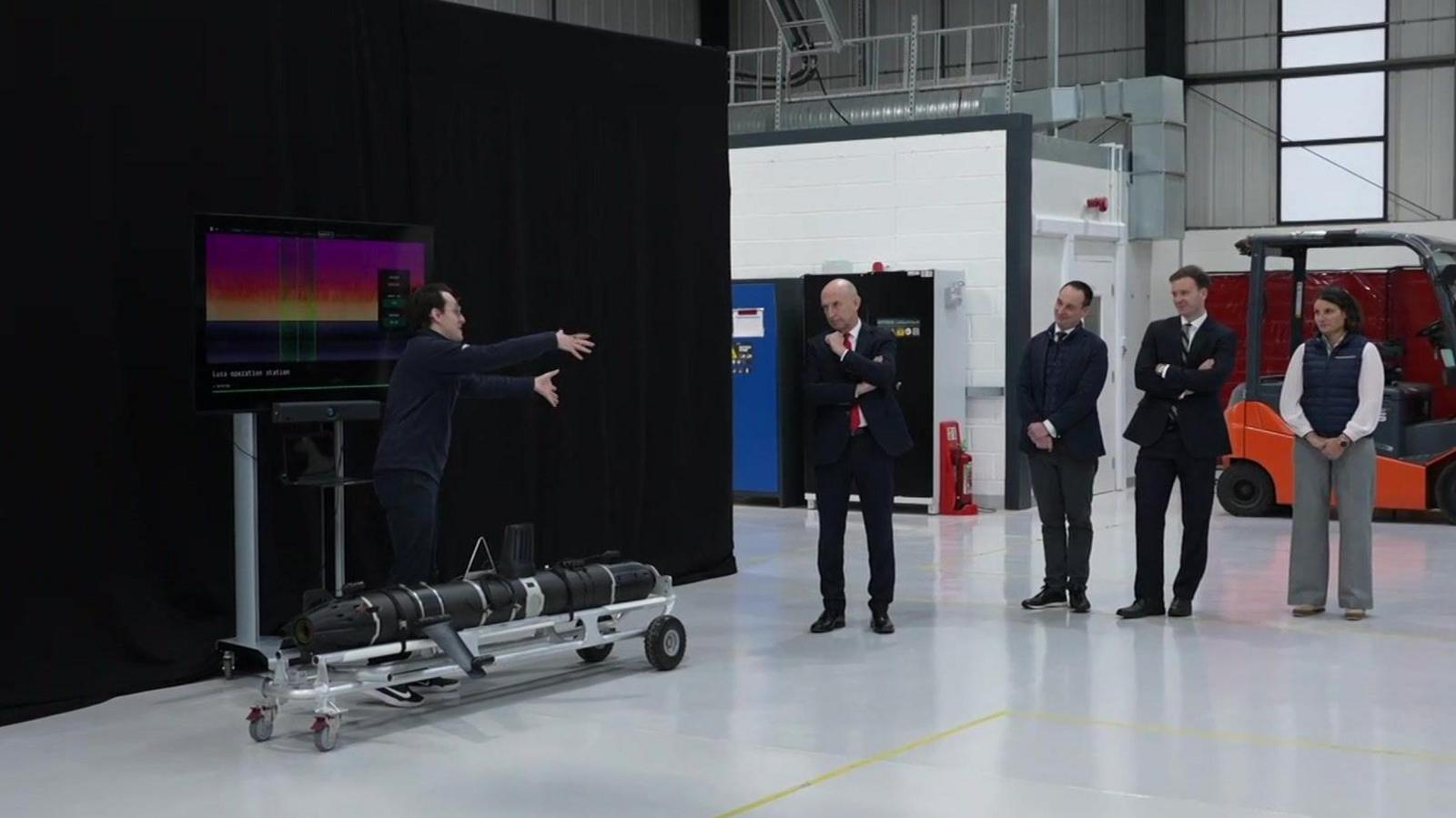 A man explains to Defence Secretary John Healey how a piece of technology, which is a long black machine on a trolley, works. Three other people are stood next to the cabinet minister in a factory.