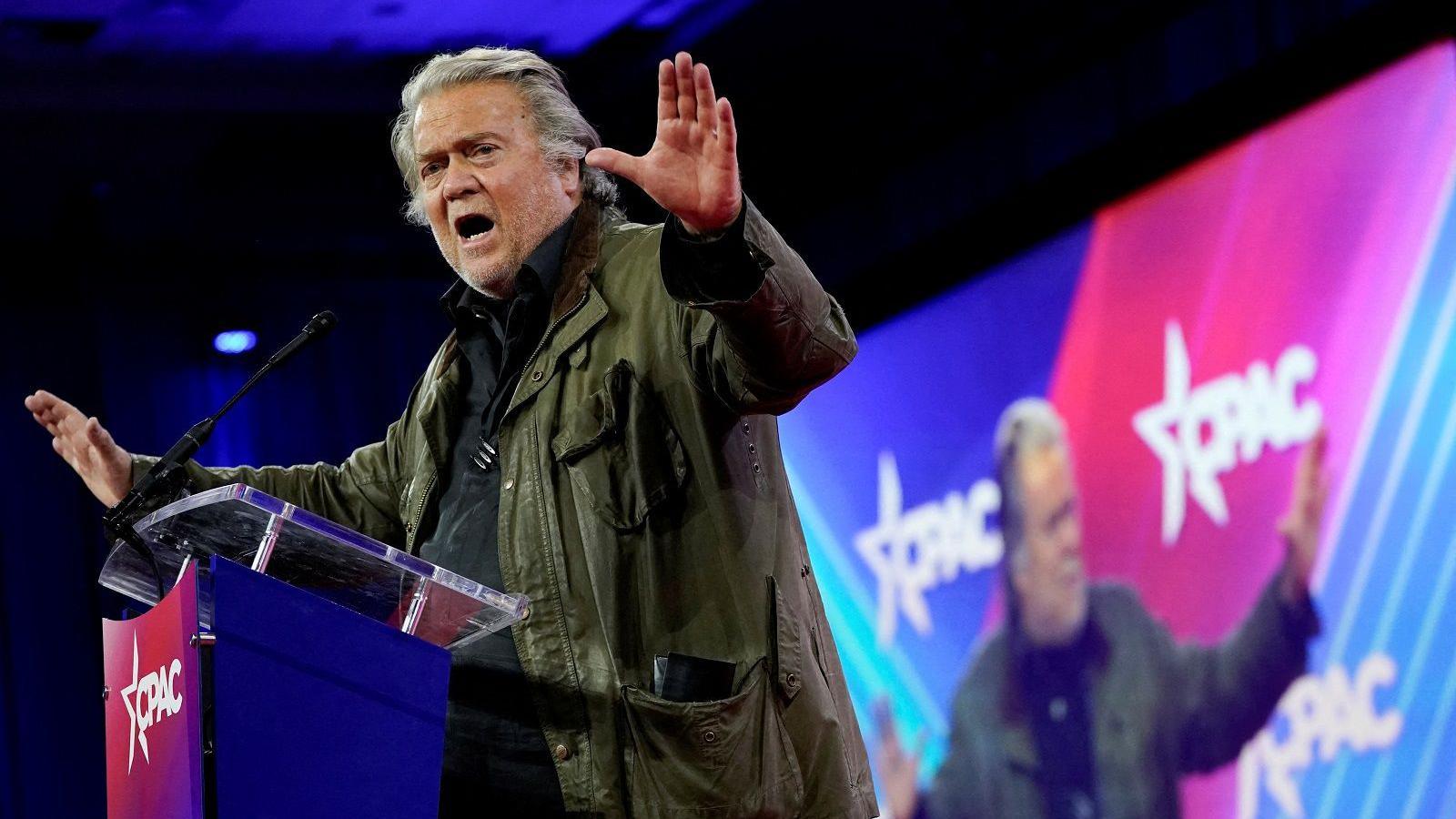 Steve Bannon in the midst of a speech in front of a large illuminated screen, waving his arms