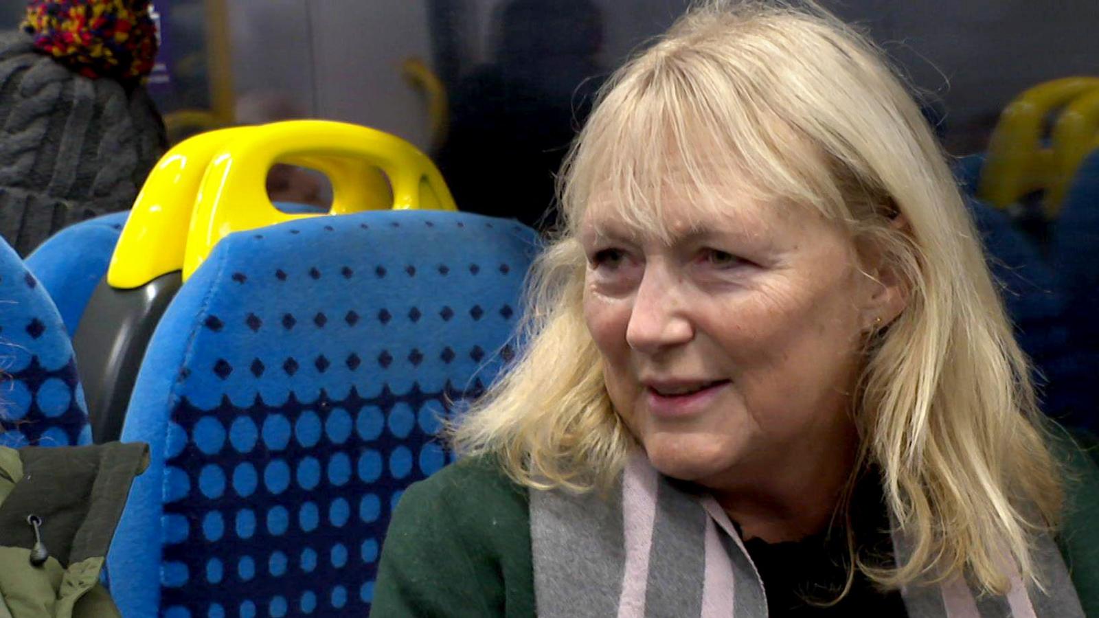 Clare Harland, a woman with blonde hair wearing a green coat, sits on a train