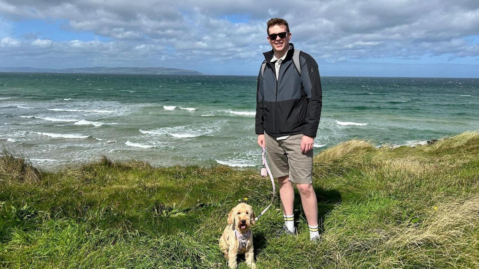 Scott Adams wearing grey raincoat and shorts, standing on grass in front of ocean with his dog. 