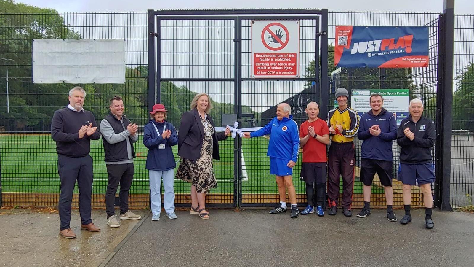 A group of people stood in front of a fence and gates at an astroturf football pitch. There are nine people, dressed mostly in football attire, but some in smart dress, and most of them are clapping.