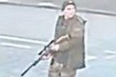 A man in camouflage gear and boots can be seen carrying what looks like a rifle with a scope across a street. He is wearing a dark hat and the image is blurry.