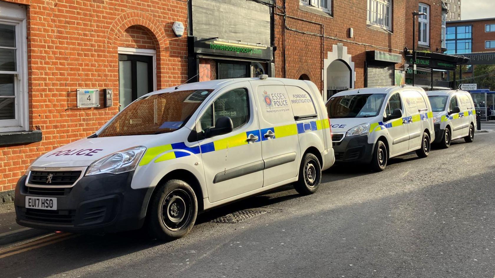 Three white forensic vans parked in a row on the street, in front of a brick building. They have Essex Police branding on them and a blue and yellow fluorescent panel.