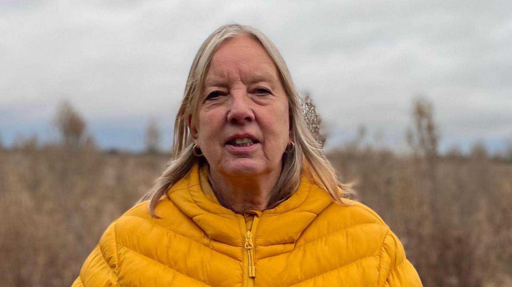 Carol Duff is looking at the camera wearing a yellow coat, she is standing in front of an overgrown field.