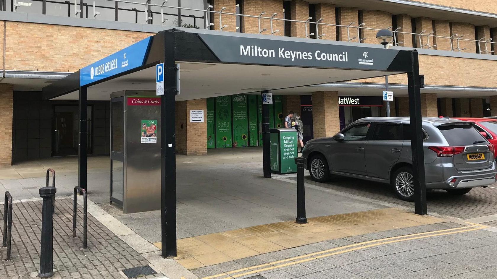 A picture of the outside of Milton Keynes City Council headquarters. The building is several stories high, with light brown bricks, with a covered entrance outside and a car park containung two vehicles.