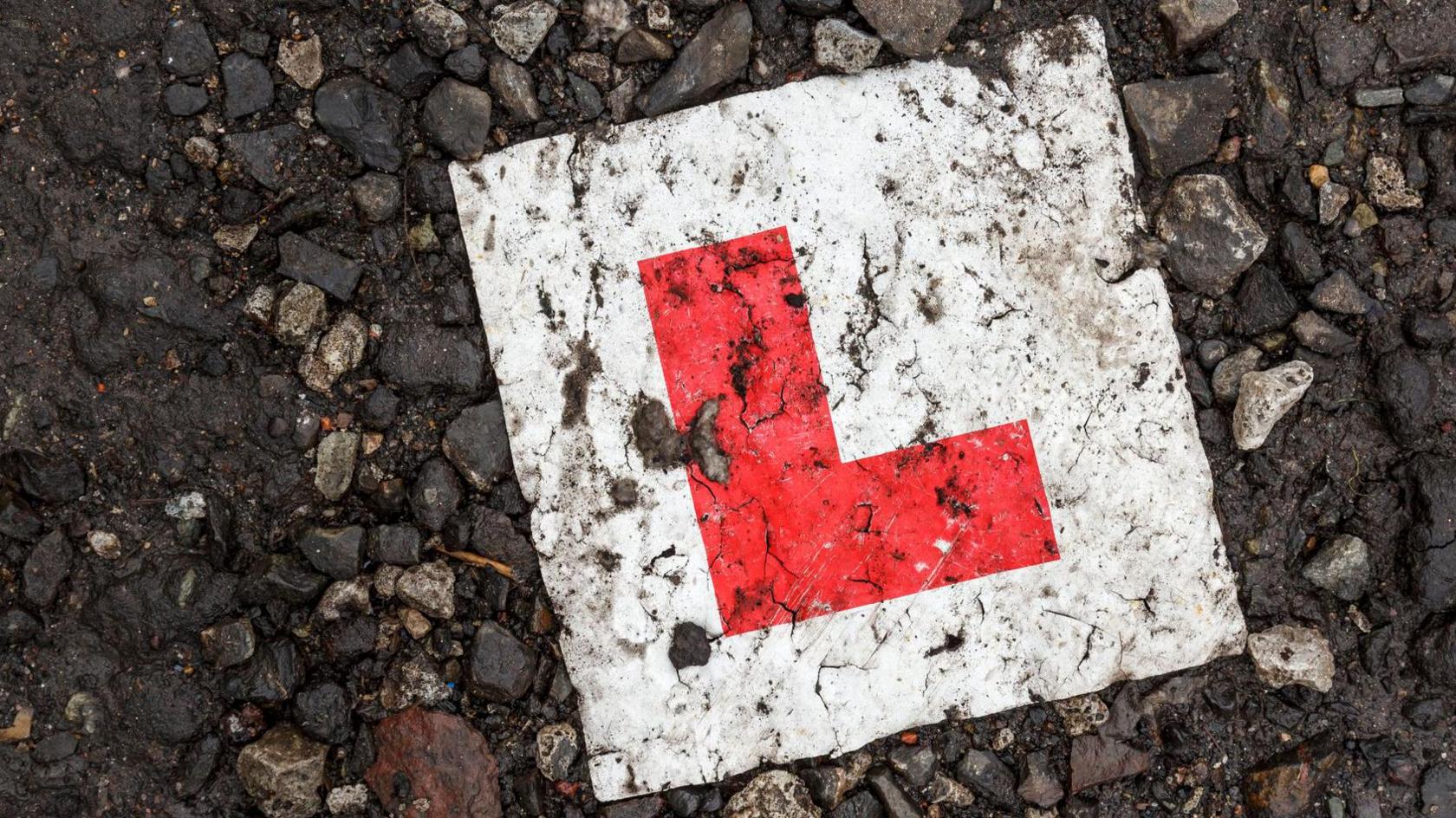 A learner plate with a red L on a white background sits on a stoney floor. 
