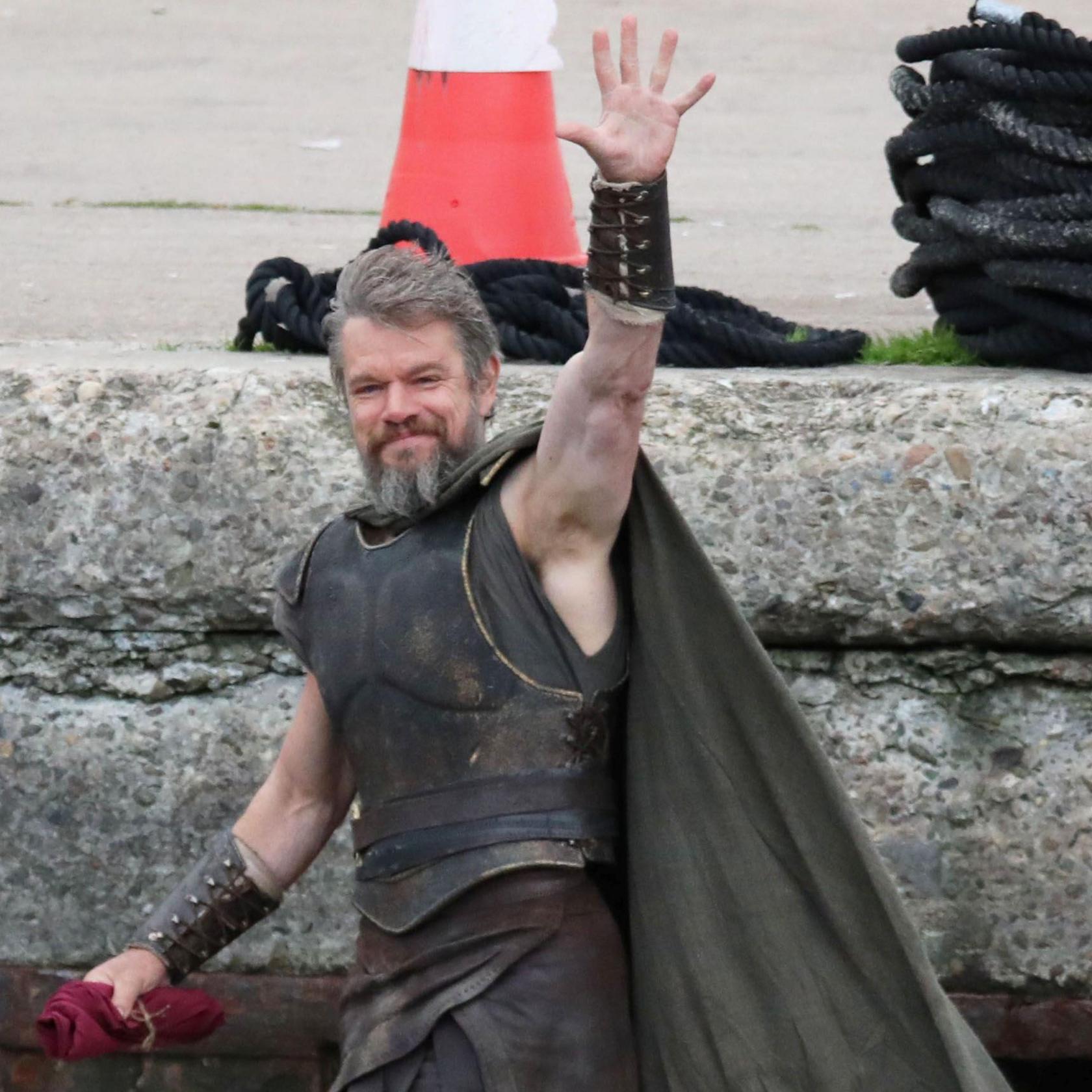 Matt Damon is pictured at Burghead harbour. He is wearing ancient Greek style armour and a grey cloak. His hair is swept to one side and he has a bushy grey beard. The actor is smiling and raises one arm to wave a hand. Behind him are large stones of a harbour wall. On top of the wall are coils of black rope, and a red and white traffic cone.