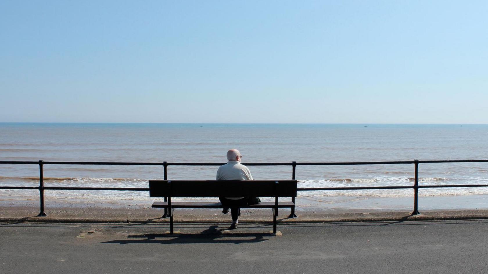 Man sat on a bench looking out to sea in Hornsea. The sky is blue and there are small waves lapping at the shore.